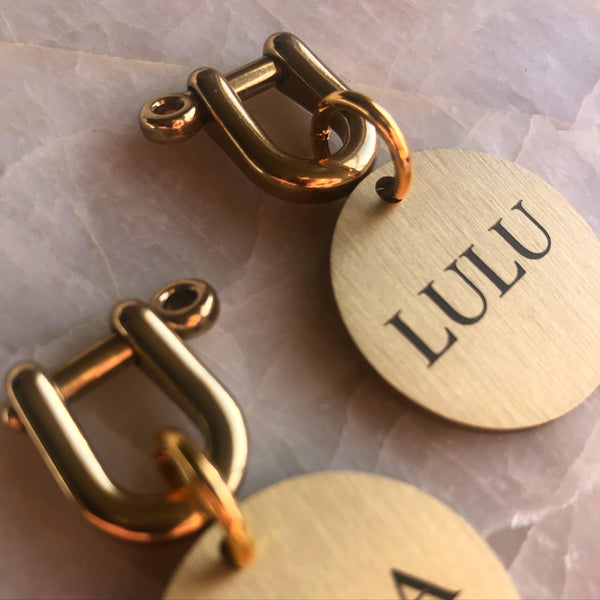 Close-up of personalized round metal tags with brushed gold finish, engraved with the name 'LULU' and a matching gold clasp, on a pale stone surface.