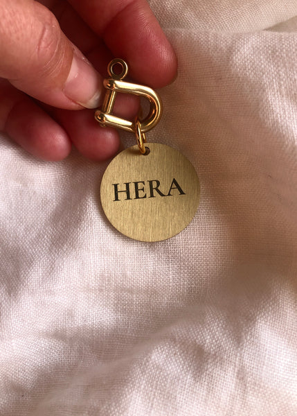 A close-up of a hand holding a gold-tone personalized pet ID tag with a D-ring clasp and a round brushed metal disc engraved with the name 'HERA', set against a soft white textured fabric background.